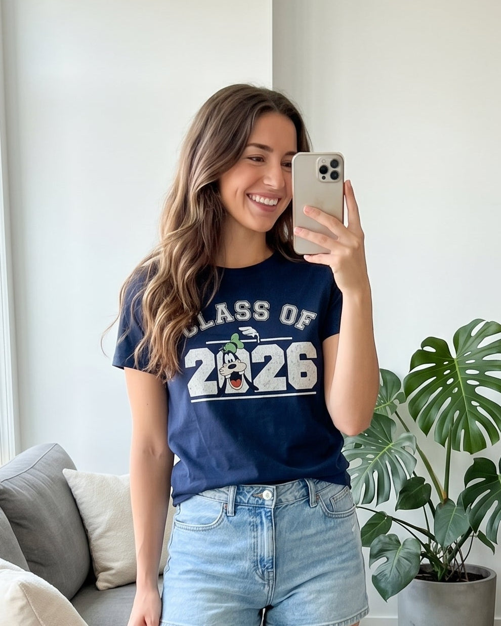Woman taking a mirror selfie wearing a 'Class of 2026' shirt in a bright room with a plant and sofa.